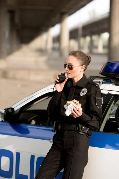 Policewoman in sunglasses talking on portable radio and holding burger near car — Stock Photo