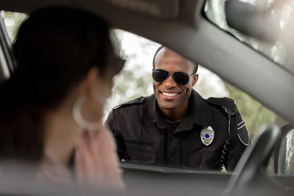 Policier afro-américain en lunettes de soleil souriant à une jeune femme assise dans une voiture — Photo de stock