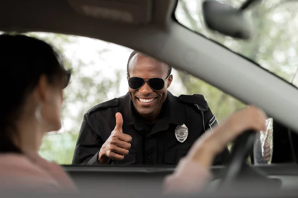 Smiling african american policeman doing thumb up gesture to young woman sitting in car — Stock Photo