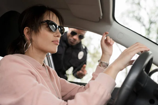 African american policeman talking to young female driver sitting in car — Stock Photo