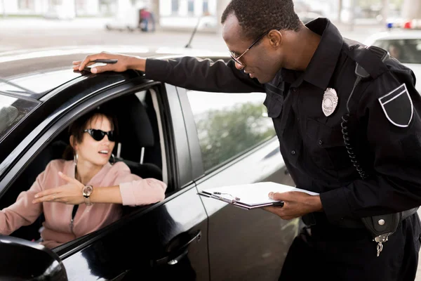 African american policeman with clipboard talking to young woman sitting in car — Stock Photo