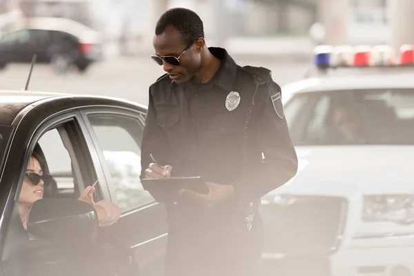 Jovem mulher no carro dando carteira de motorista para o policial afro-americano com área de transferência e caneta — Fotografia de Stock