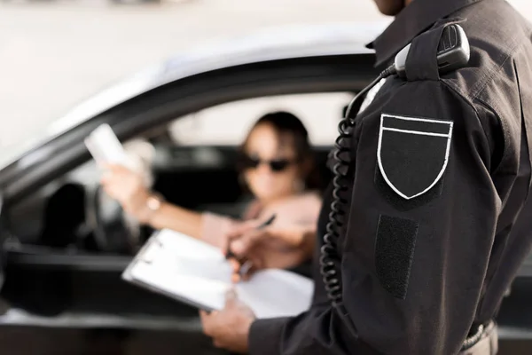 Recortado disparo de policía escribiendo en portapapeles y hablando con la mujer sentada en el coche - foto de stock