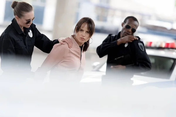 African american policeman talking on radio set while his partner arresting young woman — Stock Photo