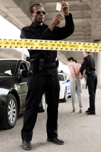 Young african american policeman in sunglasses holding plastic zipper with drugs while his partner arresting woman — Stock Photo