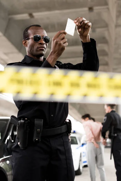 Policeman in sunglasses holding plastic zipper with drugs while his partner arresting female criminal — Stock Photo