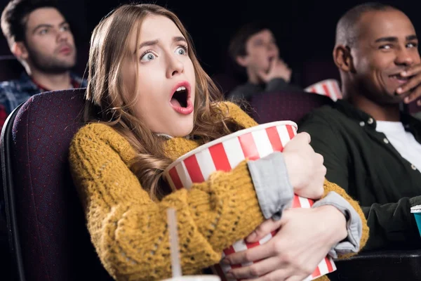 Young shocked girl holding big basket of popcorn and watching movie in cinema — Stock Photo