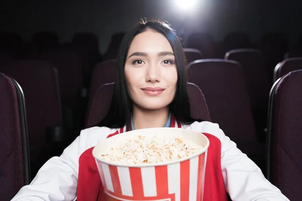 Joven asiático chica con Grande cesta de palomitas de maíz viendo película en cine - foto de stock