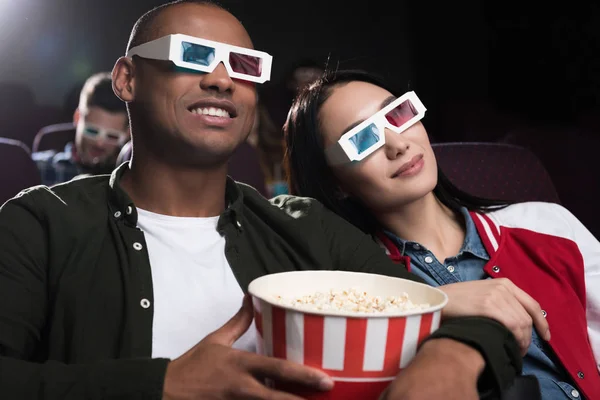 Young interracial couple in 3d glasses with popcorn watching movie in cinema — Stock Photo