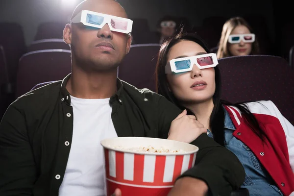 Young interracial couple in 3d glasses with popcorn watching movie in cinema — Stock Photo