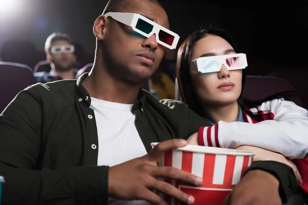 Young interracial couple in 3d glasses with popcorn watching movie in cinema — Stock Photo