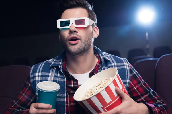 Эмоциональный красивый мужчина в 3D очках с попкорном и содовой, смотрящий кино в кино — стоковое фото