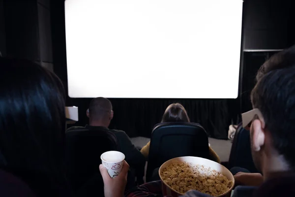 Vista posteriore di amici con popcorn e bevande seduti al cinema con schermo bianco vuoto — Foto stock