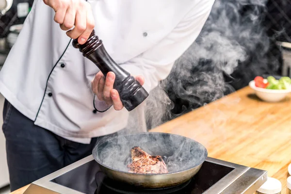 Cropped image of chef seasoning frying meat with pepper — Stock Photo