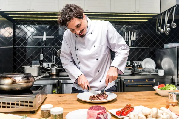 Feliz chef cortando carne frita con cuchillo y tenedor - foto de stock