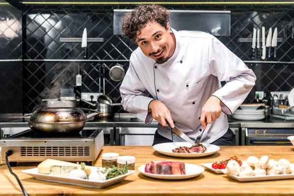 Chef sonriente cortando carne frita con cuchillo y tenedor - foto de stock