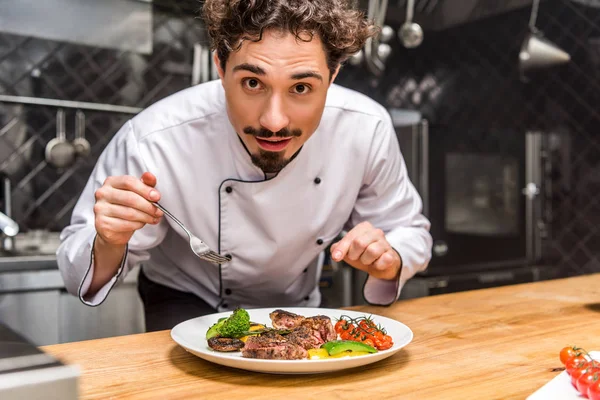 Guapo chef de pie con tenedor sobre verduras cocidas con carne - foto de stock