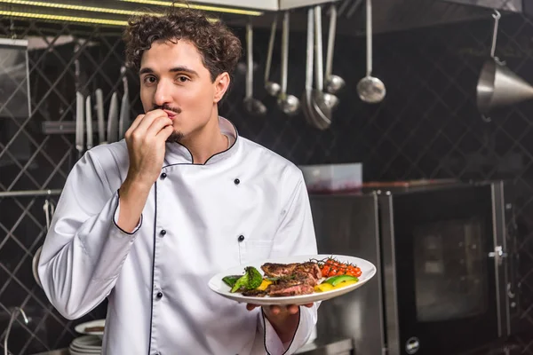 Chef in possesso di piatto con verdure fritte e carne e mostrando delizioso segno — Foto stock