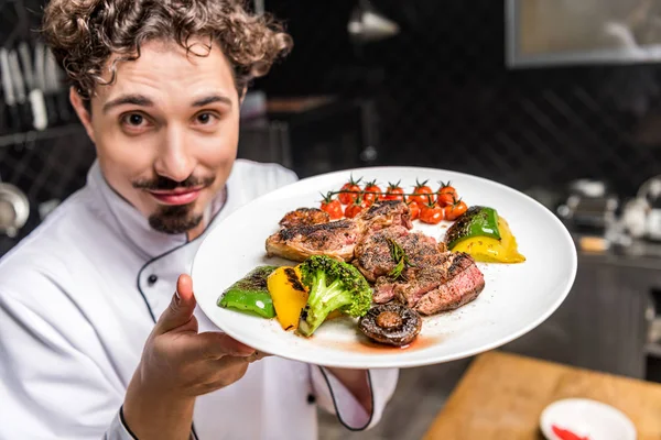 Улыбающийся шеф-повар показывает приготовленные овощи с мясом на тарелке — стоковое фото