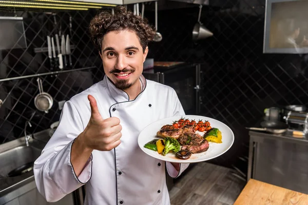 Щасливий шеф-кухар показує великий палець і тримає варені овочі з м'ясом — стокове фото