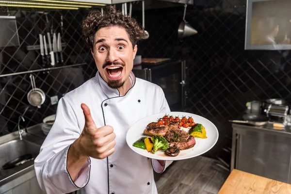 Kreischend lächelnder Koch zeigt Daumen hoch und hält gekochtes Gemüse mit Fleisch in der Hand — Stockfoto