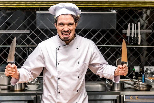 Chef haciendo muecas y sosteniendo dos cuchillos en las manos - foto de stock