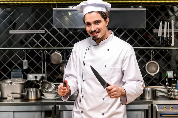 Chef souriant debout avec des couteaux dans la cuisine — Photo de stock