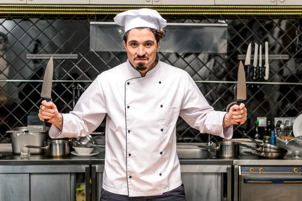 Молодой шеф-повар гримасничает и держит ножи — стоковое фото