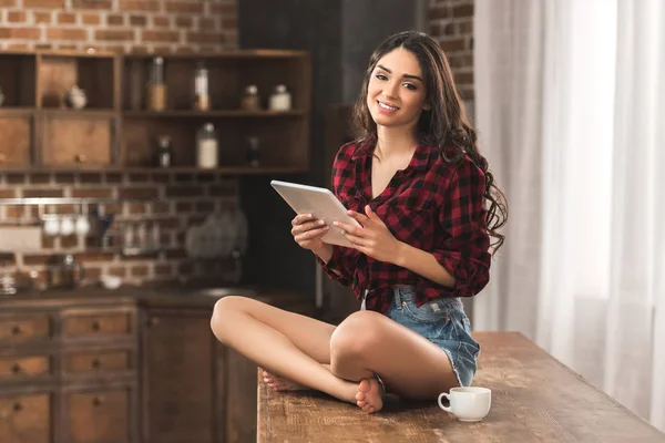 Chica con camisa a cuadros y pantalones cortos de mezclilla sonriendo a la cámara mientras usa la tableta digital en casa - foto de stock