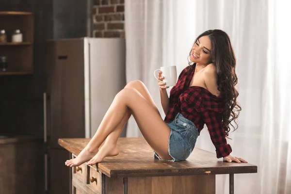 Sexy girl in checkered shirt and denim shorts drinking coffee while sitting on table at home — Stock Photo
