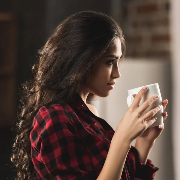 Side view of beautiful girl in checkered shirt holding cup of coffee and looking away at home — Stock Photo