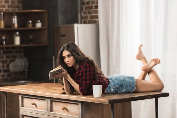Sensual jovem com livro deitado na mesa com xícara de café — Fotografia de Stock
