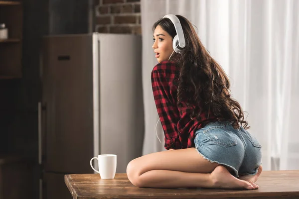 Chica sorprendida en pantalones cortos de mezclilla y auriculares mirando hacia otro lado mientras está sentado en la mesa con una taza de café — Stock Photo