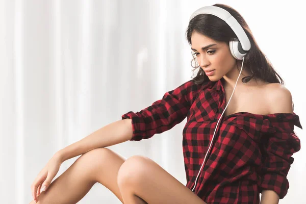 Pensive sexy girl in checkered shirt listening music in headphones at home — Stock Photo