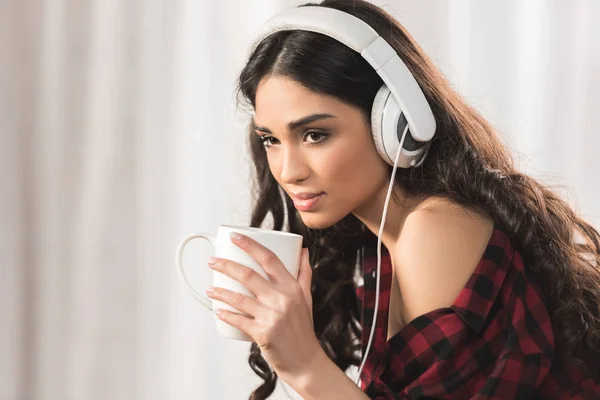 Atractiva joven escuchando música en auriculares y tomando café en casa — Stock Photo