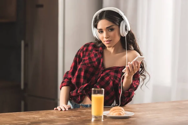 Chica con camisa a cuadros con auriculares y el uso de teléfono inteligente mientras desayuna en casa - foto de stock