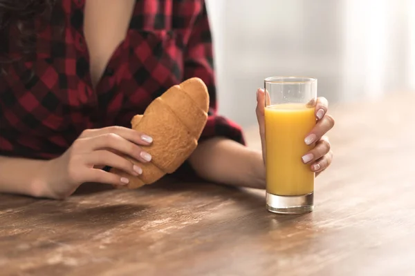 Cropped shot of girl in checkered shirt holding croissant and glass of orange juice for breakfast — Stock Photo