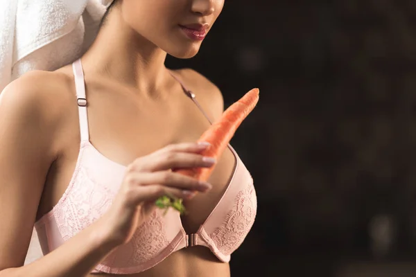 Обрезанный снимок сексуальной молодой женщины в лифчике и полотенце, держащей морковь на черном — стоковое фото