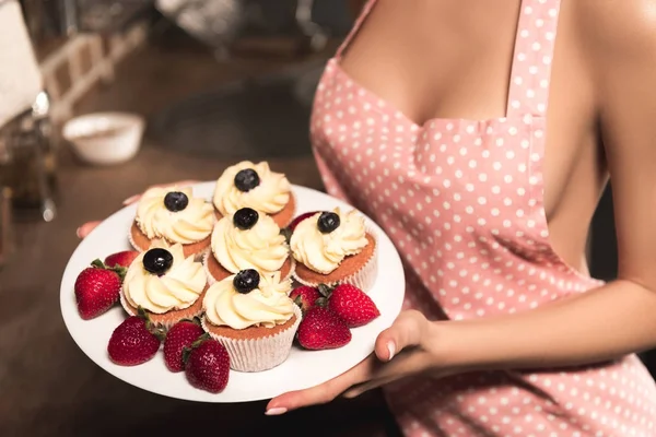 Recortado disparo de sexy chica en delantal celebración plato con cupcakes y fresas - foto de stock