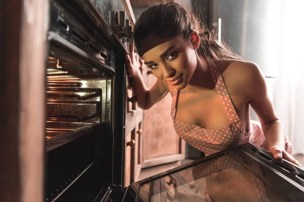 Sexy joven mujer en delantal abriendo horno y sonriendo a la cámara - foto de stock