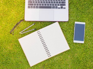 Yeşil çimenlerin üzerinde Laptop, defter, akıllı telefon ve gözlük Üstten Görünüm