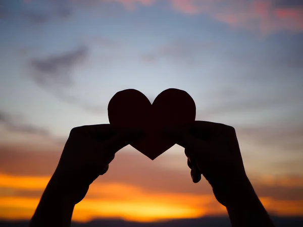 Silhouette Hand hebt das rote Papierherz mit verschwommenem Sonnenlicht während des Sonnenuntergangs, — Stockfoto