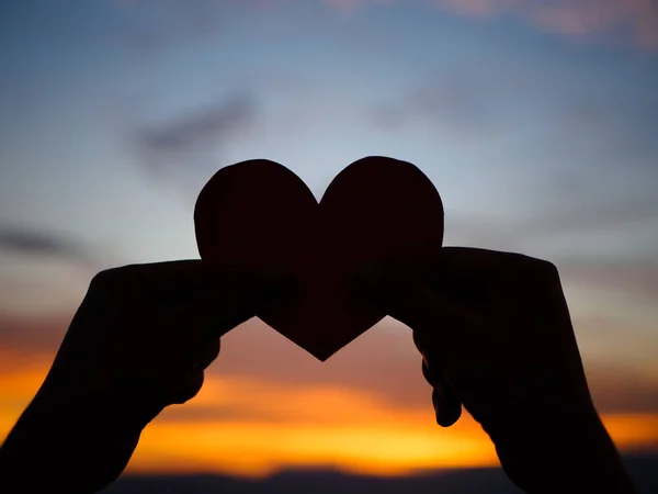 Silueta mano está levantando el corazón de papel rojo con la luz del sol borrosa durante la puesta del sol, día de San Valentín — Foto de Stock