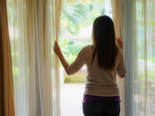 Achteraanzicht van een jonge vrouw met dat de gordijnen open om te kijken uit het grote licht raam thuis — Stockfoto