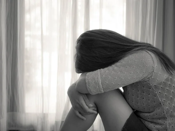 Чорно-біла сумна жінка обіймає коліно і плаче. Сумна жінка сидить наодинці у порожній кімнаті біля вікна або дверей — стокове фото