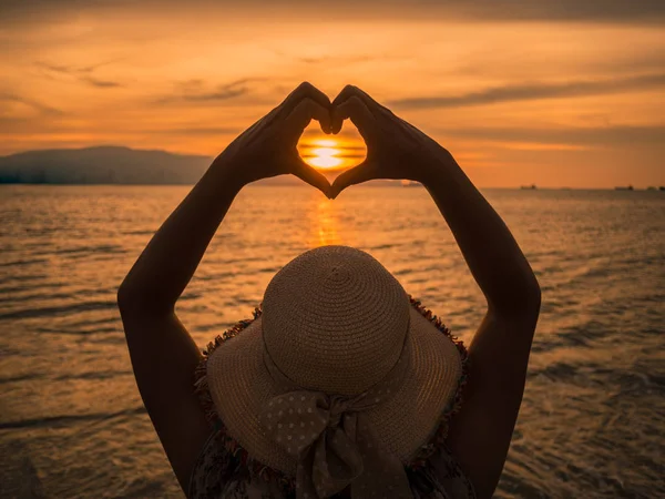 Молодая девушка, держась за руки в форме сердца, обрамляет закат солнца на берегу океана — стоковое фото