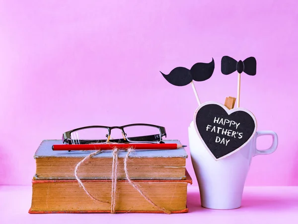 Il concetto della festa del papà. Felice padre giorno testo su pila di libri, regali, tazza di tè con baffi neri su sfondo rosa — Foto Stock