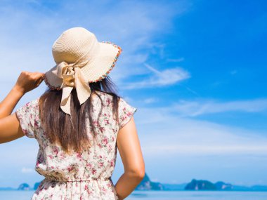 Yaz tatilindeki kadın hasır şapka ve plaj elbisesi giyip okyanus manzarasının tadını çıkarıyor.