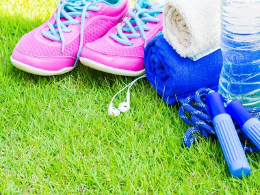 Çift pembe spor ayakkabı, havlu, kulaklık, su şişesi ve yeşil çim sahada ip atlama. İçinde arka plan orman veya park iz. Spor çalıştırmak için aksesuarlar.