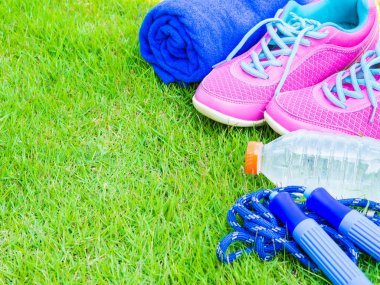 Çift pembe spor ayakkabı, havlu, kulaklık, su şişesi ve yeşil çim sahada ip atlama. İçinde arka plan orman veya park iz. Spor çalıştırmak için aksesuarlar.